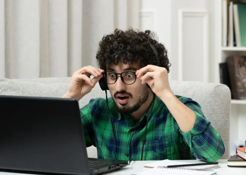Ein Mann mit lockigem Haar und Brille, der ein grün kariertes Hemd trägt, sieht überrascht aus, während er mit Laptop, Notizbuch und Stift an einem Tisch sitzt, sein Headset einstellt und auf den Bildschirm starrt - vielleicht recherchiert er über Physio Lübeck.