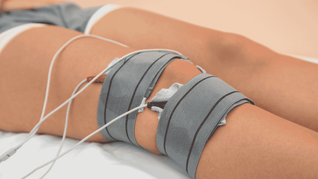Nahaufnahme des Knies einer Person mit zwei grauen Elektrodenpads, die mit Drähten für die Elektrotherapie während einer Physio Lübeck-Sitzung verbunden sind, während der Patient auf einer weißen Fläche ruht.