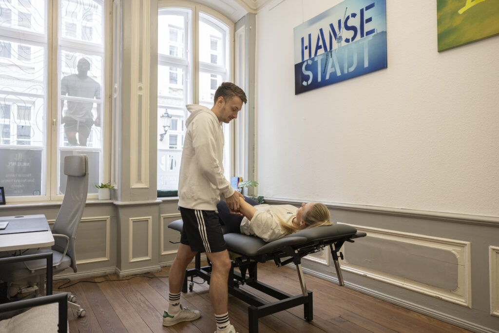 Ein Physiotherapeut im weißen Kapuzenpulli untersucht das Knie einer Frau, die auf einer Behandlungsliege in einem hellen, mit großen Fenstern und modernem Dekor ausgestatteten Klinikraum von Physio Lübeck liegt.