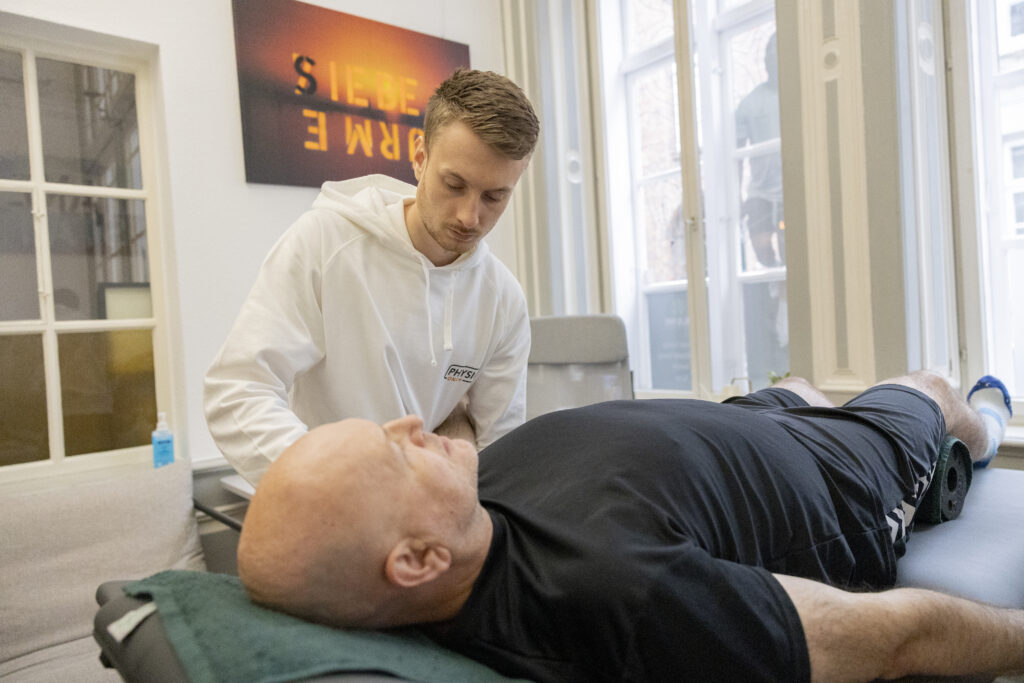 Ein Therapeut im weißen Kapuzenpulli untersucht einen glatzköpfigen Mann, der auf einer Behandlungsliege in einer hellen, modernen Klinik für Physiotherapie Lübeck mit großen Fenstern liegt.